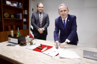 dois homens de terno posam para a foto, um dos quais assina um documento sobre a mesa