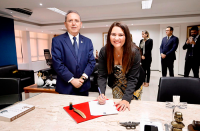 mulher assina documento sobre a mesa. Ao seu lado, homem de terno posa para a foto 