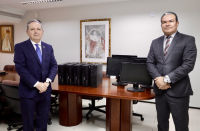 Dois homens de terno posam para a foto. Ao meio, equipamentos informáticos exibidos sobre a mesa