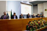 mesa de honra com oito homens sentados. Arranjo de flores na frente da mesa. Ao fundo, imagem de  data show e bandeiras hasteadas.