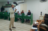 Foto de um homem branco em pé numa sala de paredes brancas, falando ao microfone, rodeado de pessoas sentadas em carteiras escolares verdes.