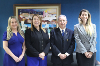 Foto de três mulheres e um homem com as mãos cruzadas à frente de seus corpos. Ao fundo, parede azul marinho com um quadro de arte colorida.