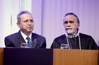 dois homens, o primeiro de terno e gravata azul, o segundo de toga, sentados à mesa de um plenário