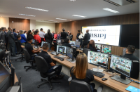 Pessoas sentadas em frente a vários monitores de imagem e ao fundo grupo de pessoas que acompanham a inauguração do centro de videomonitoramento.