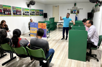 Foto de um homem branco em pé numa sala de paredes brancas com cartazes coloridos, falando para pessoas à sua volta, sentadas em carteiras escolares verdes.