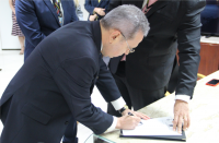 Foto de um homem reclinado, assinando um documento apoiado sobre uma mesa de tampo claro