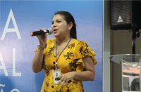 Foto de mulher branca usando vestido amarelo estampado e falando ao microfone. Ao fundo, parede azul e caixa de som preta.