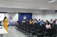 Foto de mulher usando vestido amarelo, de costas, falando ao microfone para pessoas sentadas em cadeiras de auditório.