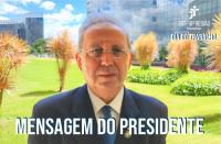 Ao fundo, gramado e plantas que decoram um jardim. À frente, homem vestido com terno e gravata em tons escuros, usando óculos de lentes arredondadas. Texto escrito de cor branca. 