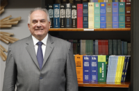 Foto de homem de pele clara e grisalho, usando terno cinza, camisa branca e gravata roxa. Ele posa para a foto, sorrindo e posicionado à frente de uma estante de madeira, na qual se veem vários livros de capas coloridas nas prateleiras.