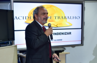Foto de homem branco com cabelos e barba grisalhos, de perfil, usando terno escuro e falando ao microfone, na frente de uma tela de datashow mostrando um capacete amarelo e o texto ACIDENTE DE TRABALHO em letras azuis.