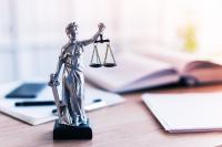 Estatua da Justiça sobre a mesa. Ao fundo, estão desfocados um livro aberto, papeis de anotação e uma caneta