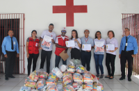 Parede de fundo na cor branca e uma cruz centralizada na cor vermelha, porta de madeira com grades ao lado esquerdo. Nove pessoas, homens e mulheres, posando para a foto com o cartaz da campanha em mãos. À frente, alimentos arrecadados.