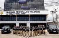 Policiais judiciais fardados, posam para foto em frente a fachada do prédio-sede do TRT-16, ladeados por duas viaturas pretas. O grupo esta perfilado em "V", tendo à frente uma mulher e apenas homens nas laterais.