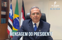 Homem sentado em uma cadeira preta, usa óculos de grau com lentes arredondadas, um terno azul escuro, uma camisa branca e uma gravata azul. Ao fundo, uma parede na cor branca, três bandeiras hasteadas e uma estante com livros à esquerda.