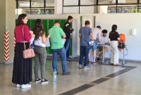 Fila com candidatas e e candidatos para entrar na sala de aplicação de prova. Homem sentado em carteira coletando as informações.