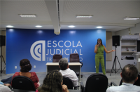 Ao fundo, uma mulher vestida com um macacão na cor verde e falando ao microfone. Ao fundo, uma caixa de som preta e uma parede azul com letras brancas, com texto escrito ESCOLA JUDICIAL. À frente, pessoas sentadas em cadeiras pretas. 