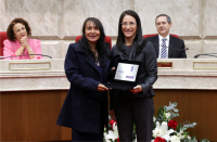 Duas mulheres posando para foto, com vestimentas formais, segurando uma placa de premiação. Atrás delas um arranjo central de flores vermelhas e brancas. Ao fundo,  uma mulher e um homem estão sentados em poltronas vermelhas, apoiados em uma mesa.