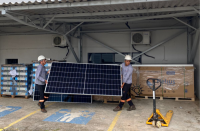 A imagem mostra dois homens usando equipamento de proteção individual, carregando painel solar em formato retangular, de cor azul escuro. À direita, um equipamento de transporte das placas solares. Ao fundo, placas empilhadas sobre pallet de madeira. 