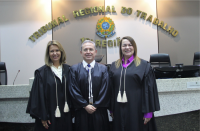 Foto de um homem claro, ladeado por duas mulheres de cabelos claros. Todos usando togas de juiz e posando para a câmera fotográfica. Atrás deles, parede com a inscrição TRIBUNAL REGIONAL DO TRABALHO em letras douradas, acima de um brasão colorido da República do Brasil.