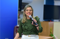 Foto de uma mulher usando uma camisa verde e uma calça branca, falando ao microfone. Ao fundo, parede azul e televisão na cor preta. 