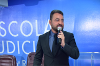 Foto de homem branco com cabelos e barba escuros, de frente, mas olhando para o seu lado esquerdo. Ele fala ao microfone e está posicionado à frente de uma parede azul com a inscrição ESCOLA JUDICIAL em letras brancas.