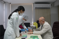 Homem usando terno claro, sentado em uma cadeira preta, apoiado com os braços em cima da mesa. À frente da mesa, duas mulheres vestidas com jalecos brancos.