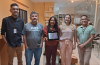 Três homens e duas mulheres posam para foto sorrindo, em uma sala com paredes na cor laranja. A mulher ao centro segura uma placa de premiação.