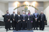 Foto de cinco homens e duas mulheres, lado a lado, posando para câmera fotográfica. Todos usam toga preta de juiz. Atrás deles, parede com a inscrição TRIBUNAL REGIONAL DO TRABALHO em letras douradas, acima de um brasão colorido da República do Brasil.