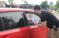 Foto de um homem claro com cabelos escuros, fardado, usando calça cáqui, camisa preta e óculos escuros. Ele segura panfletos com a mão esquerda e entrega um bottom amarelo com a mão direita ao motorista de um carro vermelho. 