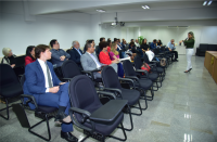 Foto de uma mulher, falando ao microfone para pessoas sentadas em cadeiras de auditório.