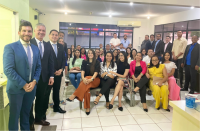 Grupo de pessoas posando para foto, alguns na parte central, sentados em cadeiras e outros, dos extremos, em pé. A foto acontece dentro de uma sala com paredes na cor branca.