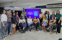 Grupo de pessoas, posando para foto, alguns na parte central sentados em cadeiras e outros em pé. Ao fundo, vê-se tela de projeção em cor predominantemente roxa, com texto na cor branca escrito "PROGRAMA TRT NA ESCOLA 2023". A foto acontece dentro de uma sala com paredes na cor branca.