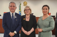 Três pessoas posando para foto, sendo um homem e duas mulheres. Ambos estão com as mão cruzadas à frente de seus corpos. O homem, ao lado esquerdo, vestido com terno azul escuro, a mulher que está no meio está vestida com roupas de cor preta, e a da esquerda está com um vestido verde estampado.