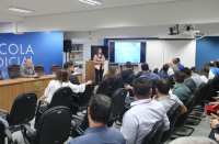 Foto de um homem e uma mulher em posição de apresentação. O homem está sentado em cadeira de auditório de cor marrom, apoiado em mesa de madeira, e a mulher está segurando um microfone e olhando para a plateia. Ao fundo, parede azul e uma tela de datashow. A plateia está sentada em cadeiras de auditório, aparecendo todas as pessoas de costas para foto.