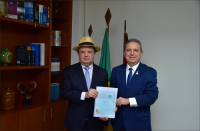 À frente, dois homens posando para a foto segurando uma cartilha de boas ações. Ao fundo, uma parede na cor branca, três bandeiras hasteadas e uma estante com livros à esquerda. 