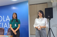 Foto de duas mulheres, a do lado esquerdo está parada observando e a do lado direito está falando e gesticulando. Ao fundo, parede azul, mesa de madeira e cadeiras na cor marrom.