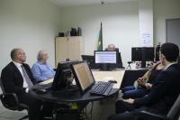 Foto realizada em uma sala de reunião de paredes brancas, uma mesa com computadores em cima e pessoas reunidas analisando uma tela de computador.