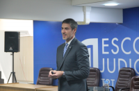 Foto de um homem que está em pé, vestindo terno escuro, em frente a uma mesa e cadeiras