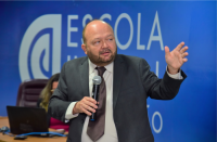 Homem vestido com terno escuro, camisa branca e gravata de cor marrom. Com a mão direita, segura o microfone e gesticula com a mão esquerda. A parede ao fundo da imagem na cor azul. 