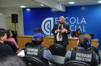 Um homem vestido com um terno escuro, uma camisa branca e uma gravata marrom, falando ao microfone. Ao fundo uma caixa de som preta e uma parede azul com letras brancas, com texto escrito ESCOLA JUDICIAL. À frente, pessoas sentadas em cadeiras pretas. 