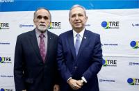 À frente, dois homens, vestidos com ternos escuros, posando para a foto. Ao fundo, backdrop branco com detalhes na cor azul, com a logomarca do TRE