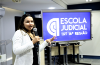 Foto de uma mulher, usando vestido preto e casaco branco, que está falando ao microfone. Ela tem cabelos compridos escuros com mechas loiras. No lado esquerdo, um telão com o nome Escola Judicial e outros equipamentos.