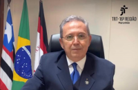 Homem sentado em uma cadeira preta, usa óculos de grau com lentes arredondadas, um terno azul escuro, uma camisa branca e uma gravata azul. Ao fundo, uma parede na cor branca, três bandeiras hasteadas.