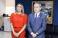 Foto de duas pessoas, um homem e uma mulher, ambos com as mãos cruzadas à frente de seus corpos. A  mulher, ao lado esquerdo, vestida com roupas de cor vermelha. O homem, vestido com terno escuro, camisa branca e gravata azul.