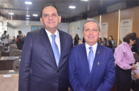 Dois homens em um plenário posando para foto, ambos vestidos com terno e gravata azul e camisa branca. O homem da direita usa óculos de grau. Ao fundo, algumas pessoas sentadas e outras em pé.
