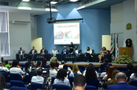 Foto de várias pessoas (homens e mulheres) sentadas num auditório participando de seminário; no destaque autoridades da solenidade de abertura. Na parede central do auditório, um projetor com as informações do evento. À direita, o brasão do Judiciário na parte superior da parede.