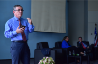 Homem em pé, vestido com camisa azul e gravata vermelha, usando óculos com lentes de grau. A mão direita ele está segurando o celular e a mão esquerda está fazendo gestos. Ao fundo, duas pessoas sentadas em poltronas de cor preta.