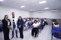 Foto tirada em uma sala de aula, com paredes brancas, cadeiras na cor azul com alunos e alunas sentados pousando para a foto. À frente, três pessoas posando para a foto, sendo duas mulheres nos extremos e um homem localizado entre elas.