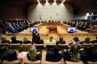 Foto de um plenário, composto por várias pessoas sentadas em poltronas de cor marrom claro. Ao centro, duas televisões. 